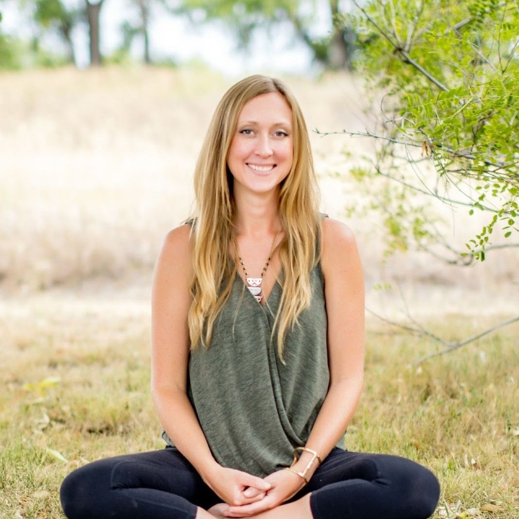 Smiling woman with long blonde hair sitting cross-legged on grass outdoors.