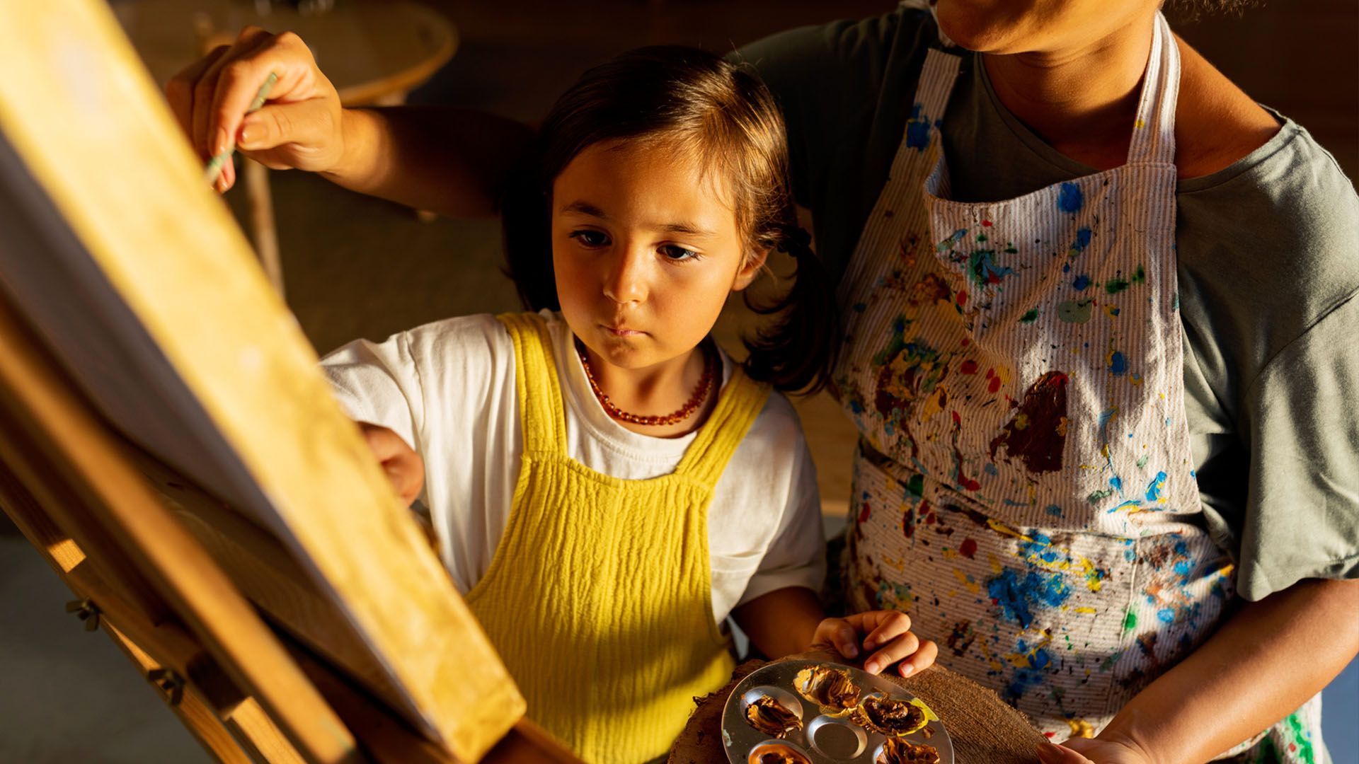 Young girl concentrating on painting on a canvas while adult holds a paint palette with mixed colors.