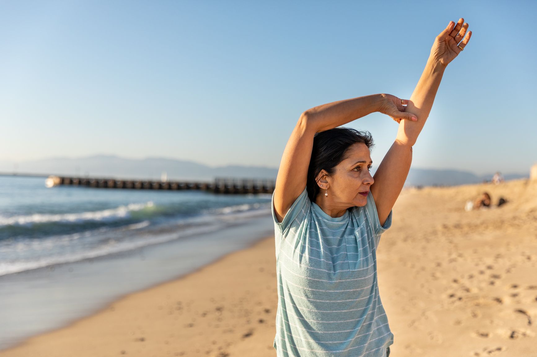Woman in a striped shirt stretching her arm on a sunny beach with ocean and pier in the background.