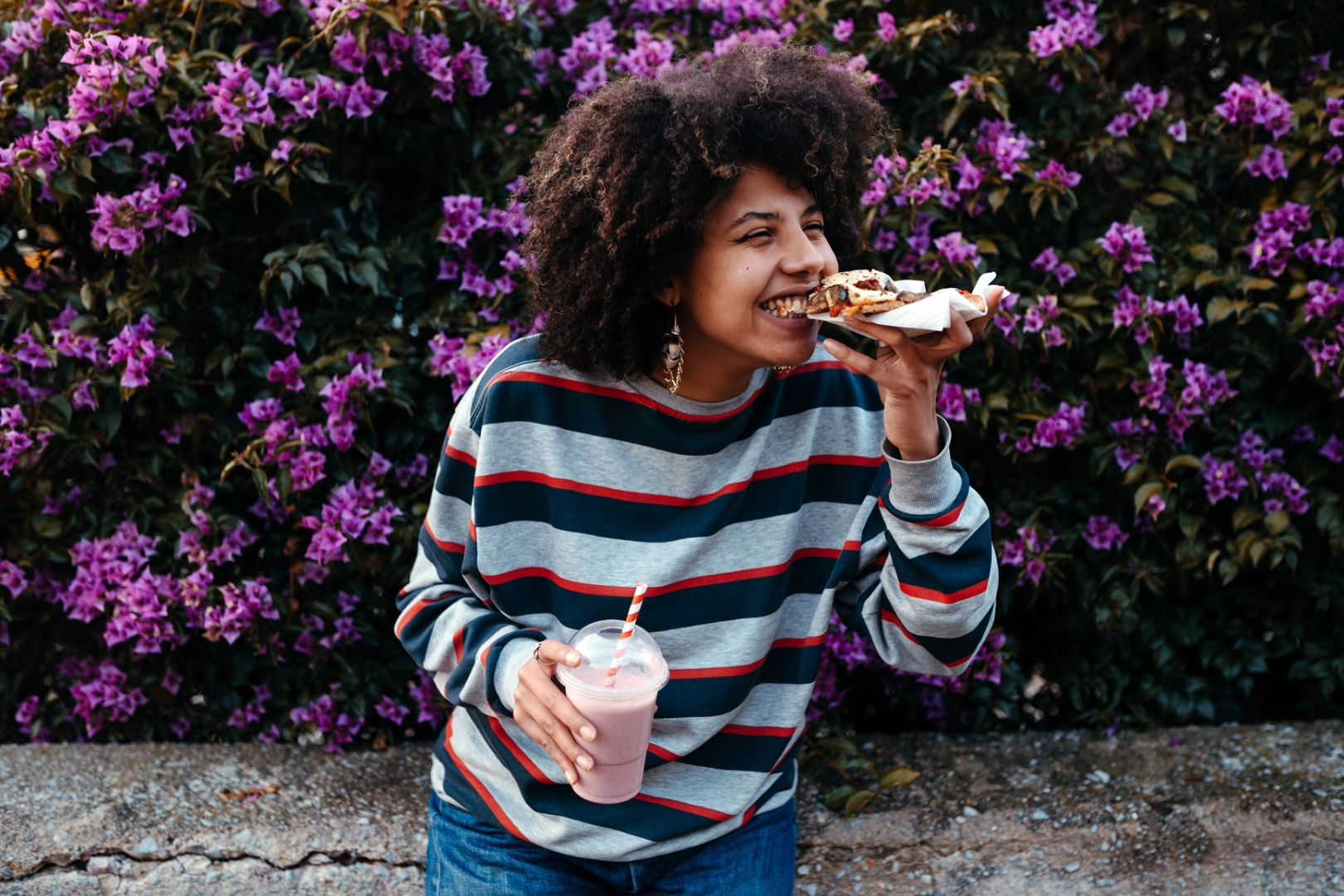 Young woman in striped sweater enjoying a slice of pizza and holding a pink smoothie with a straw in front of a purple flower bush.