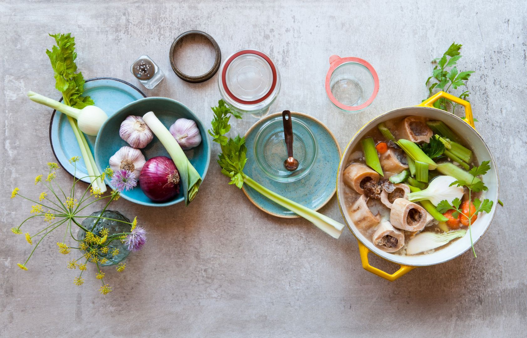 Yellow pot with bone marrow soup with vegetables including carrots, celery, onion, and herbs, surrounded by fresh garlic, onion, and celery on a blue plate.