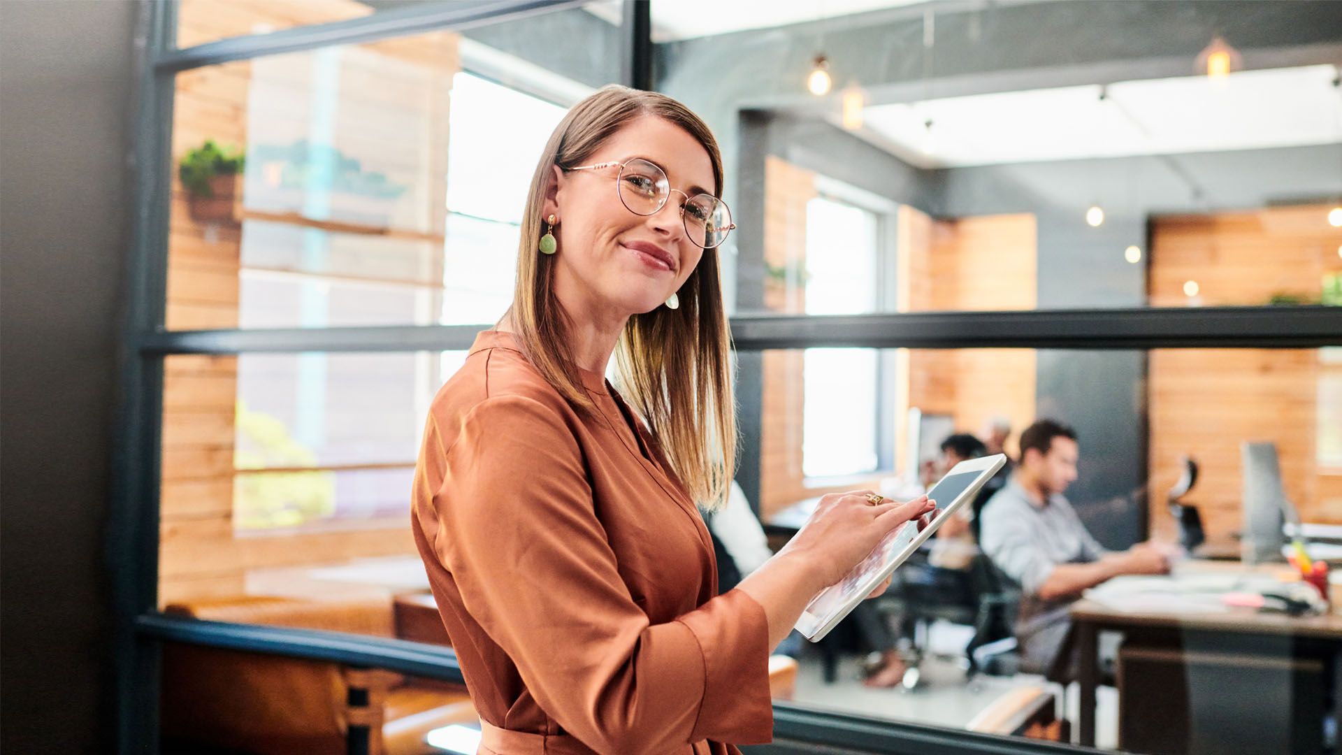 Smiling woman wearing glasses and brown blouse using a tablet in a modern office with colleagues working in the background.