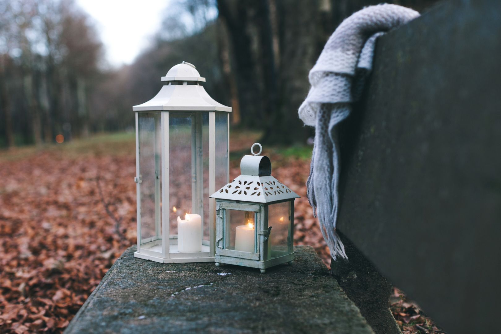 Two white lanterns with lit candles placed on a stone bench surrounded by fallen autumn leaves, with a cozy scarf draped over the bench.