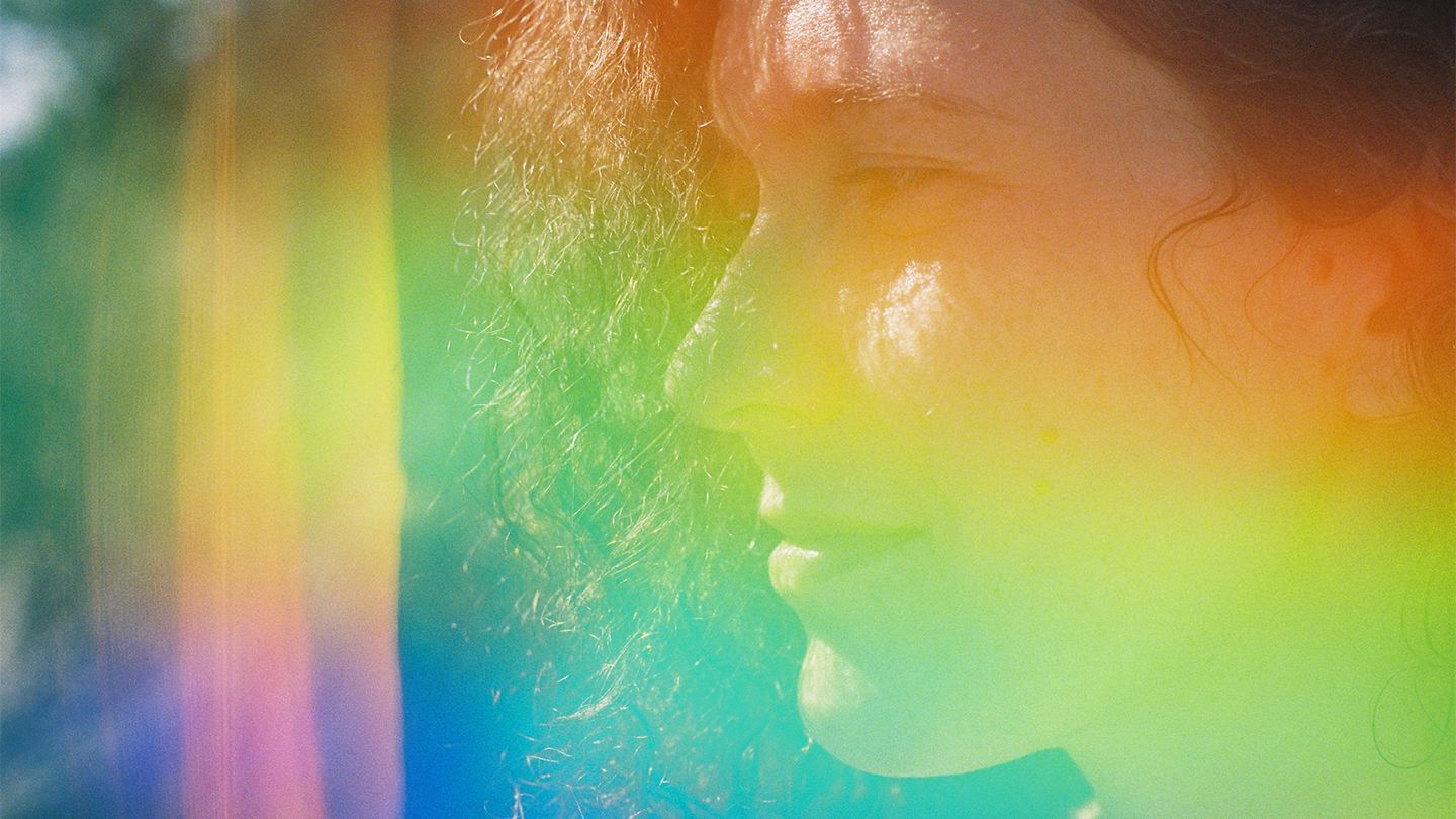 Close-up side profile of a person with curly hair, overlaid with a diffuse rainbow light effect.