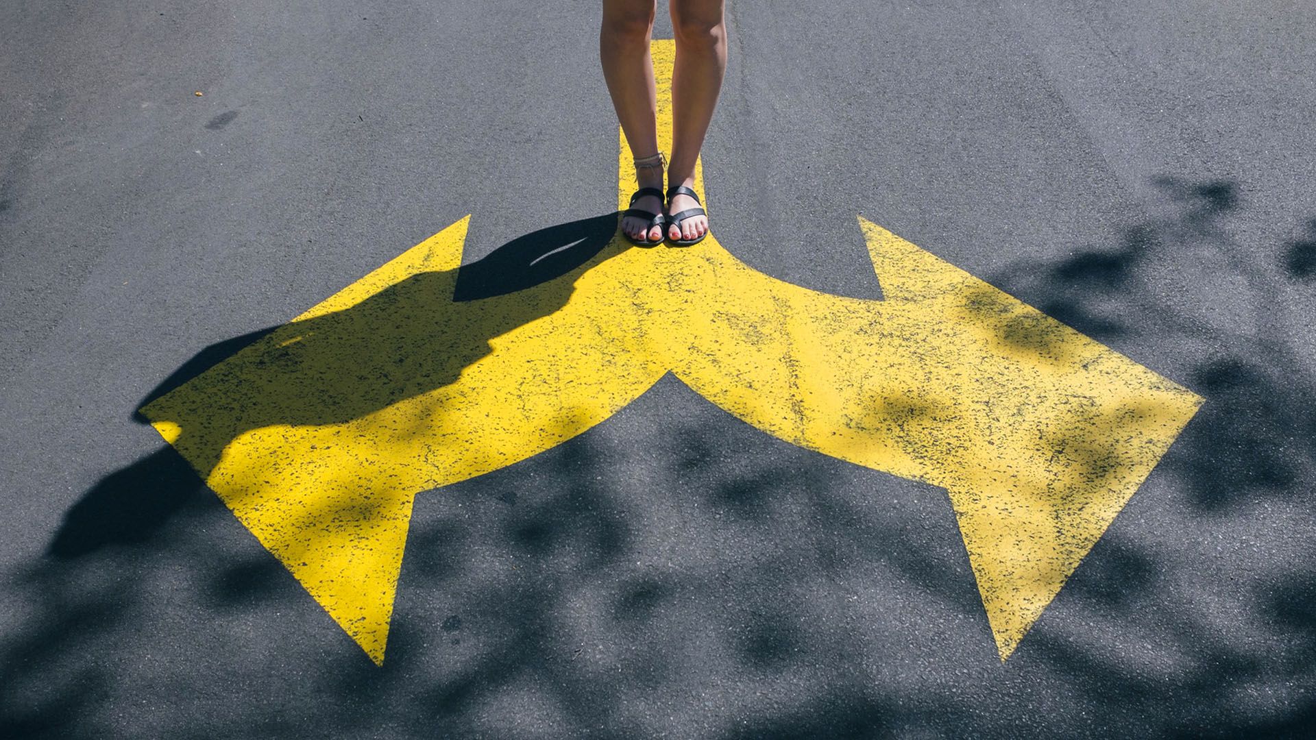 Person wearing black sandals standing on a road with large yellow arrows pointing left and right.