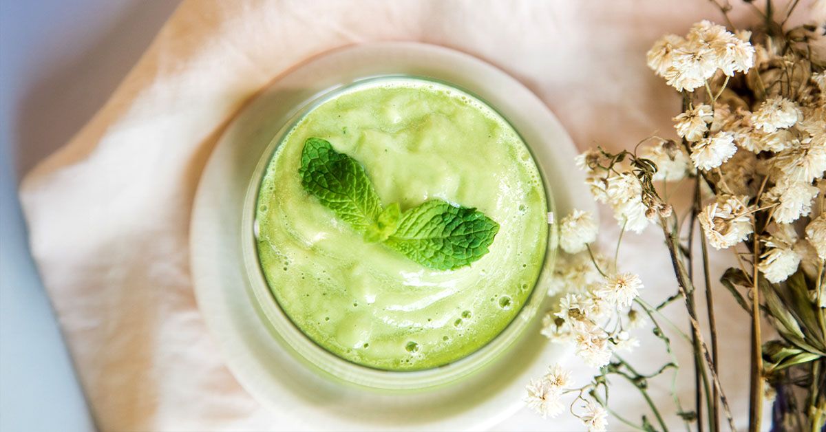 Top view of a green smoothie garnished with fresh mint leaves on a plate next to dried white flowers on a light surface.