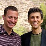 Two smiling men posing outdoors with light-colored foliage in the background.