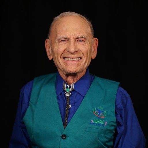 Smiling elderly man wearing a teal vest over a blue shirt with a bolo tie, against a black background.