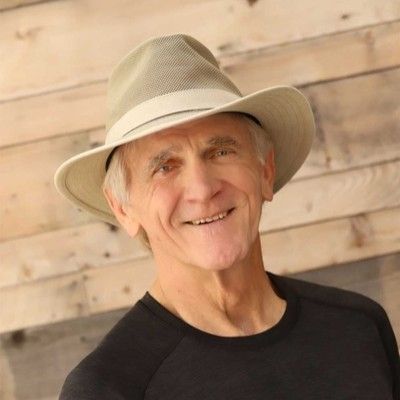 Smiling elderly man wearing a beige wide-brimmed hat and a black shirt against a wooden plank background.