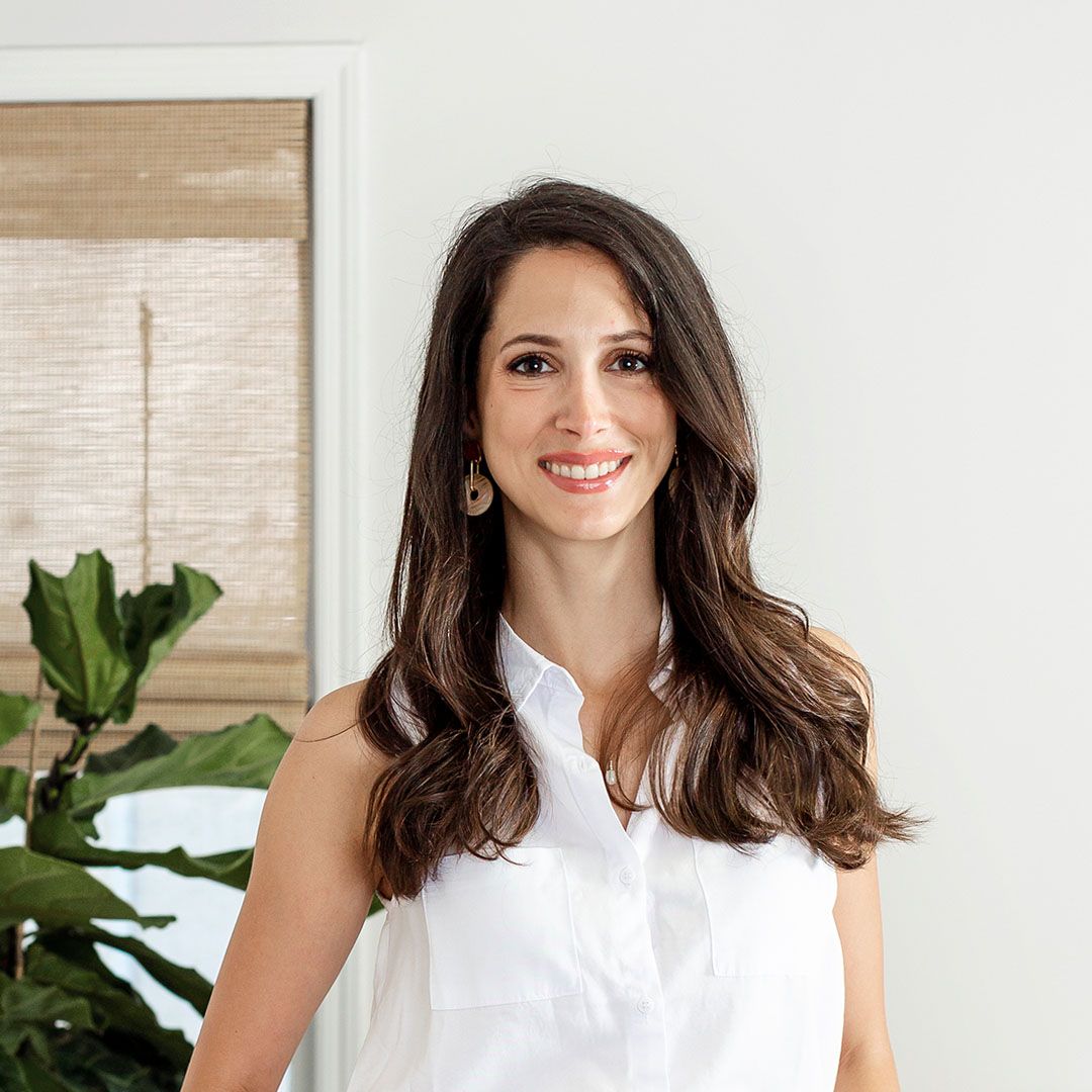 Smiling woman with long wavy brown hair wearing a sleeveless white blouse standing indoors with a plant and window blinds in the background.