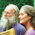Older man with long white beard and glasses playing a wooden harp beside a woman with curly hair, both in front of green foliage.