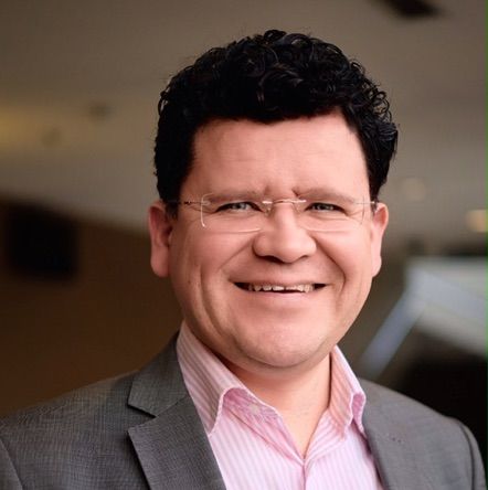 Smiling man with curly black hair wearing glasses, a gray suit, and a pink shirt.