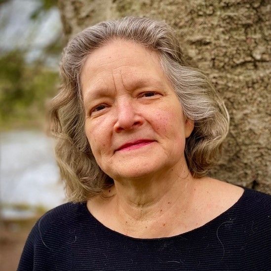 Portrait of an older woman with gray hair wearing a black top, standing outdoors near a tree.