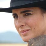 Close-up of a woman wearing a black hat with a slight smile and natural outdoor background.