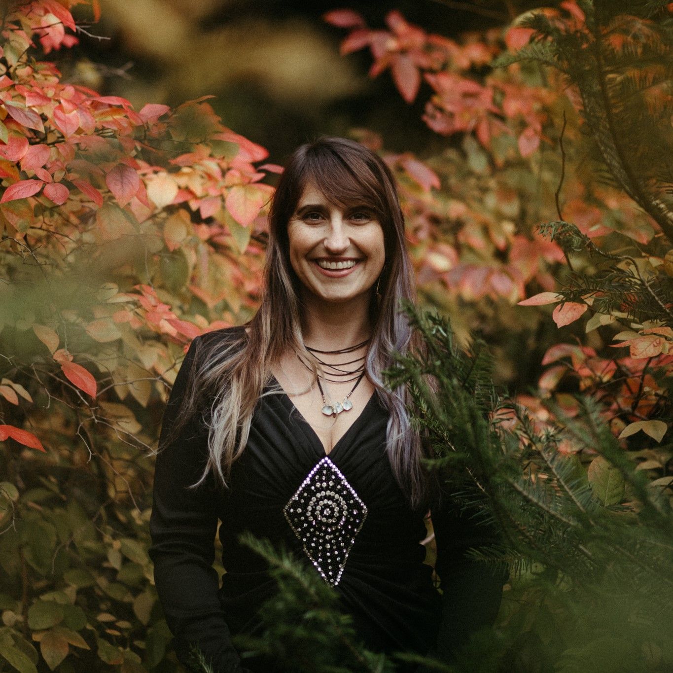 Smiling woman with long hair wearing a black dress with a sparkly diamond-shaped pattern, standing among autumnal orange and green foliage.