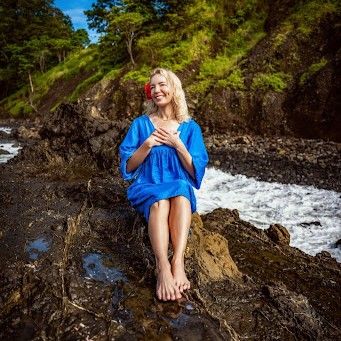 Smiling woman in a blue dress sitting barefoot on rocky shoreline with waves and greenery in the background.