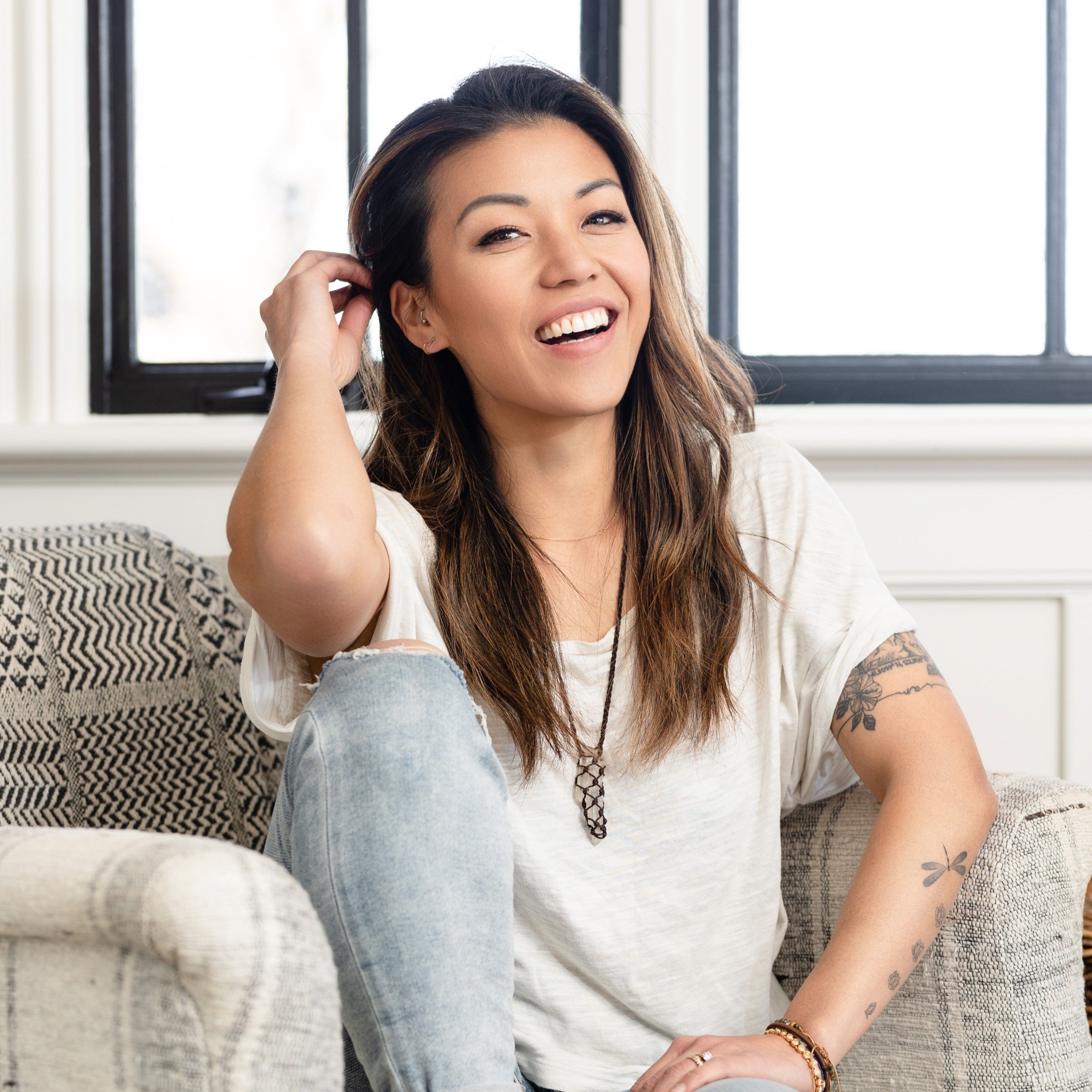 Smiling woman with long brown hair sitting on a patterned armchair, wearing a white t-shirt, ripped jeans, and displaying arm tattoos.