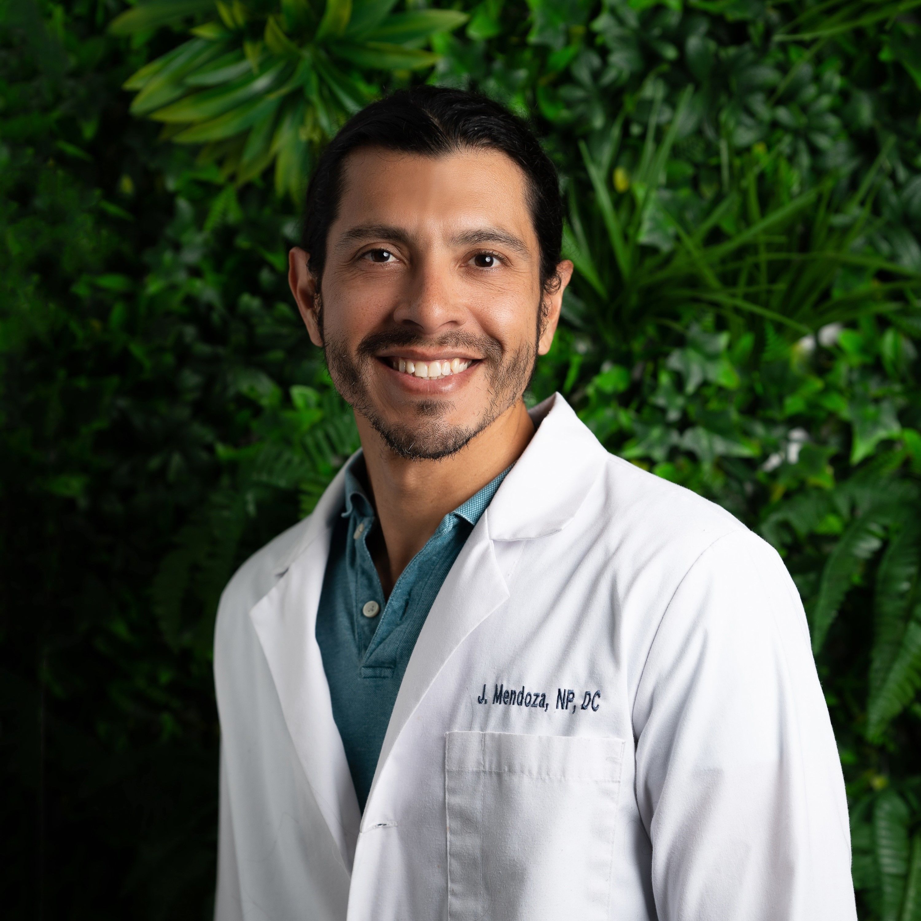 Smiling man with dark hair and beard wearing a white medical coat in front of green foliage.
