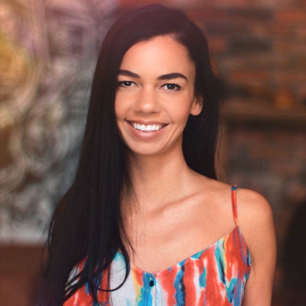 Smiling woman with long black hair wearing a colorful sleeveless top against a blurred indoor background.