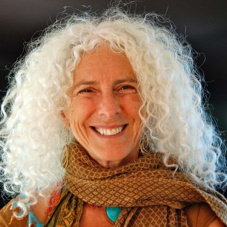 Smiling older woman with voluminous curly white hair, wearing a brown patterned scarf and gold earrings.