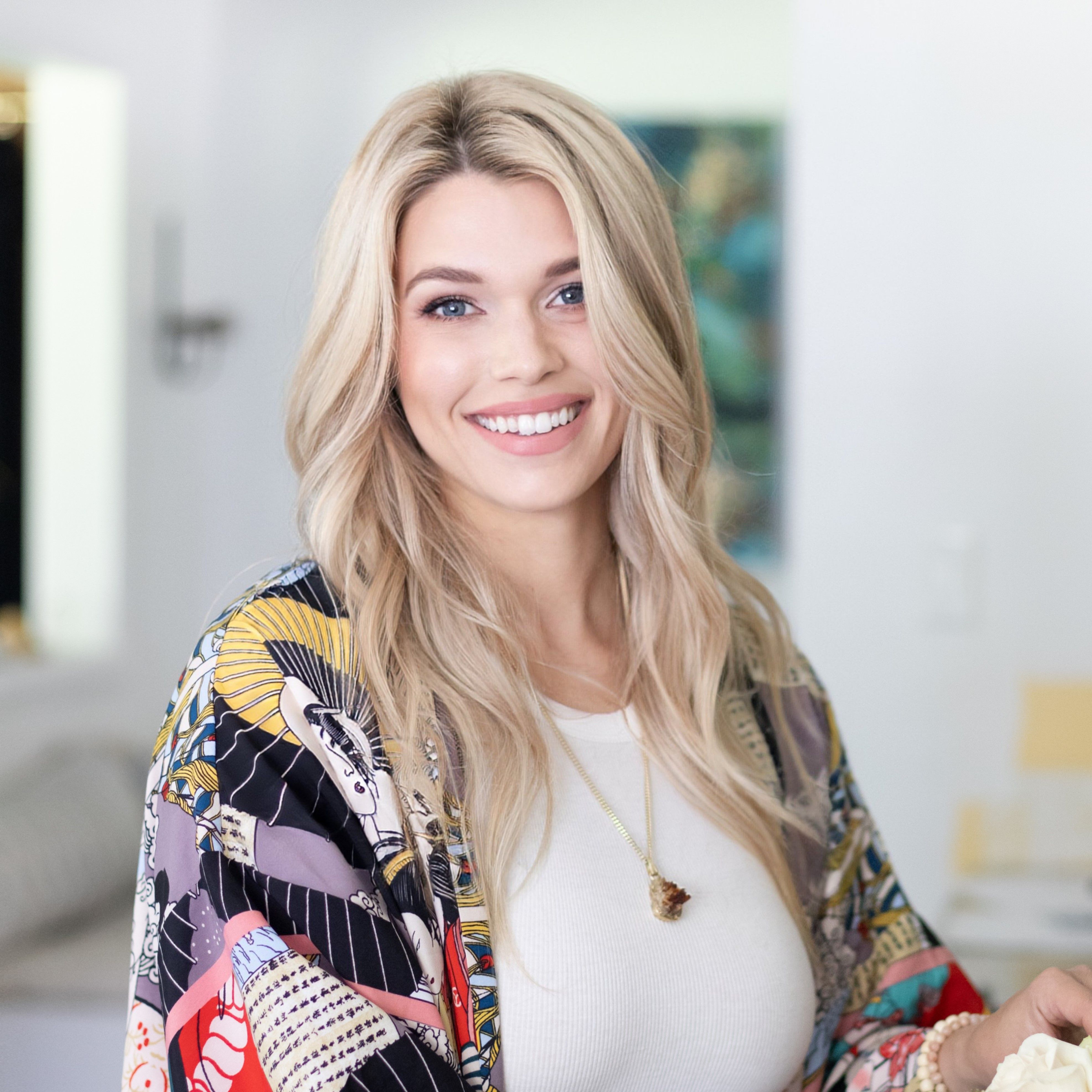 Smiling blonde woman with long hair wearing a colorful patterned jacket and white top indoors.