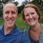 Smiling middle-aged man and woman posing outdoors with greenery in the background.