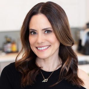 Smiling woman with shoulder-length brown hair wearing a black top and a necklace, standing in a kitchen.