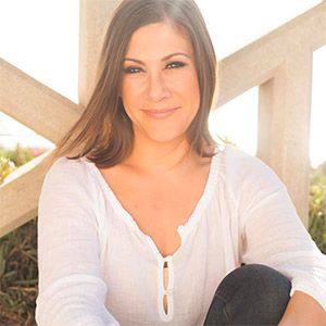 Smiling woman with long brown hair wearing a white blouse sitting outdoors against a wooden structure.