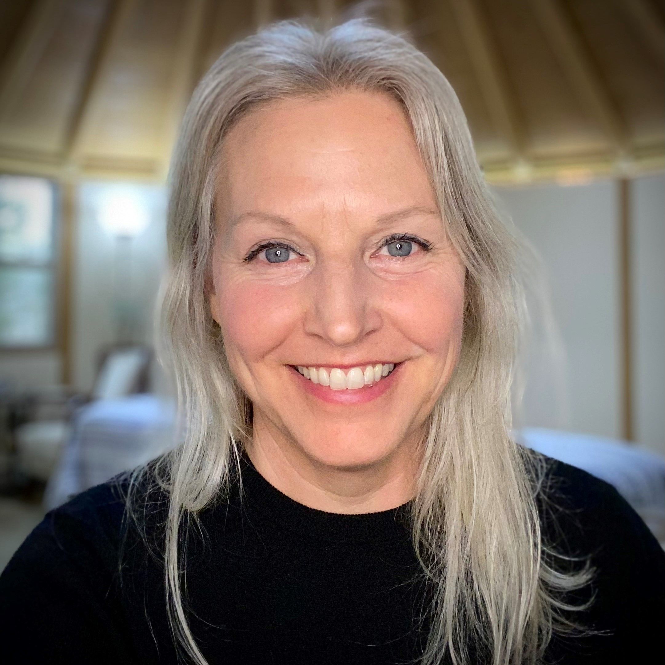 Smiling middle-aged woman with long gray hair wearing a black top in a softly lit indoor setting.
