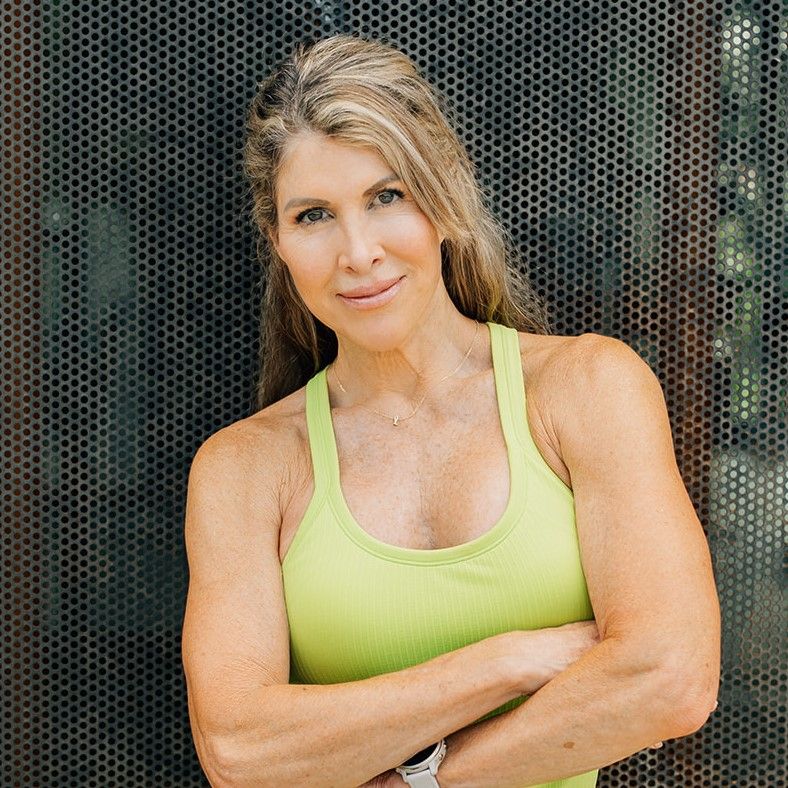 Fit woman with long blonde hair in a green tank top standing with arms crossed against a perforated metal background.