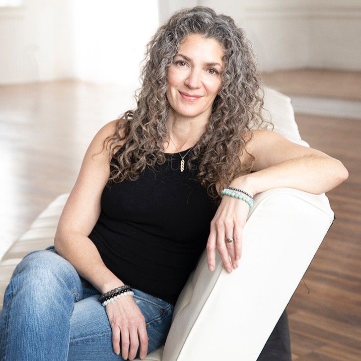 Woman with curly gray hair wearing a black sleeveless top and jeans, sitting relaxed on a white couch.