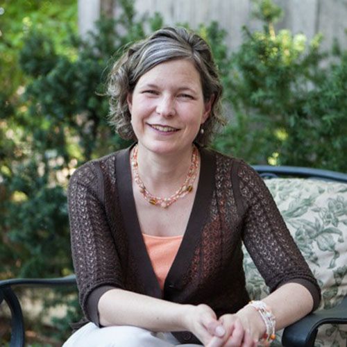 Smiling woman with short curly hair wearing a brown cardigan and a coral top, seated outdoors on a chair with greenery in the background.