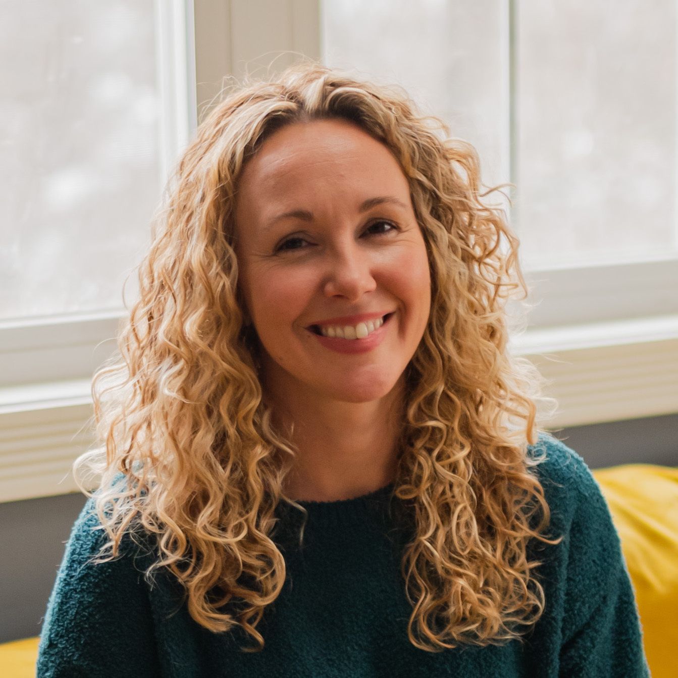 Smiling woman with long curly blonde hair wearing a dark green sweater indoors near a window.