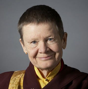 Portrait of a smiling elderly person with short hair wearing a maroon and yellow robe against a gray background.
