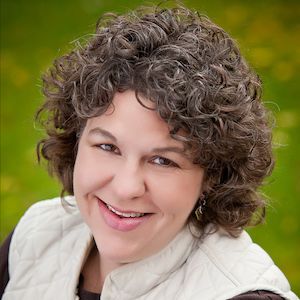 Smiling woman with curly brown hair wearing a white jacket against a green outdoor background.