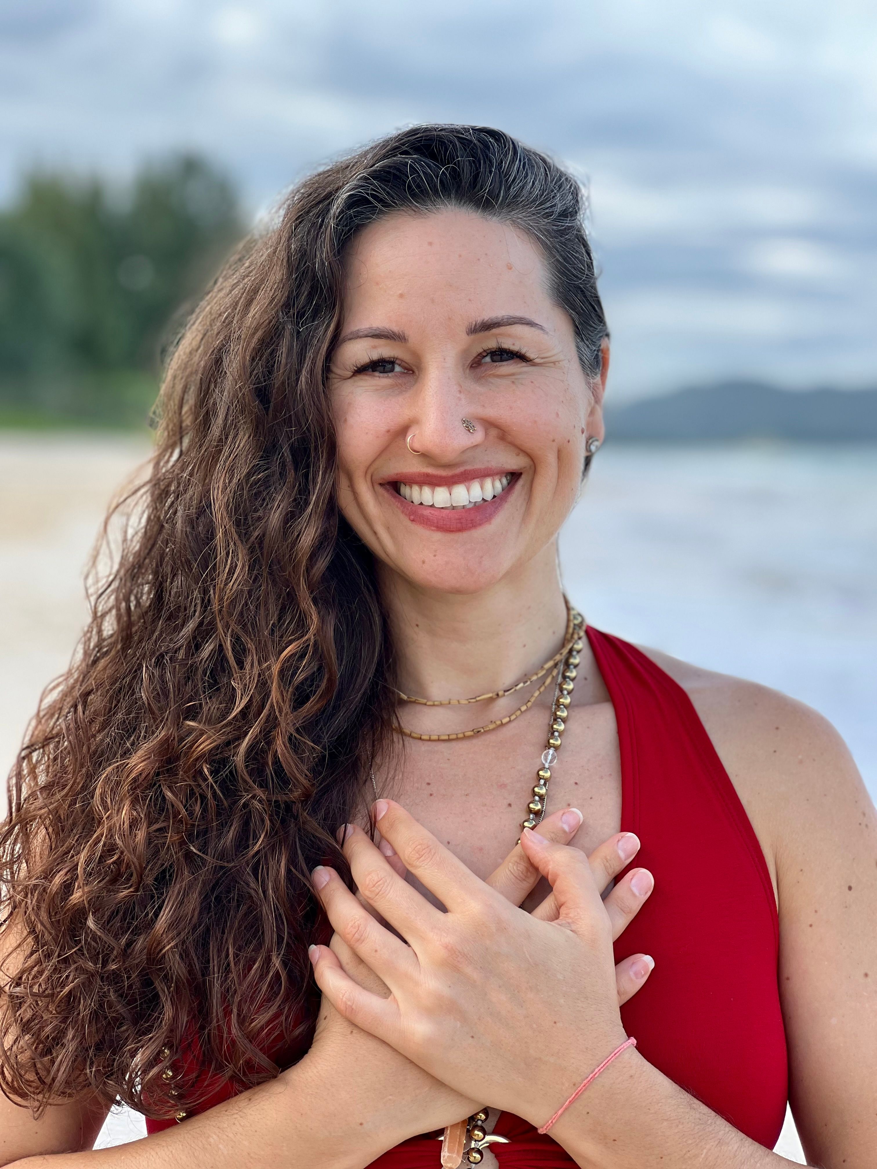 Smiling woman with long curly hair, wearing a red top and layered necklaces, standing on a beach with her hands crossed over her chest.