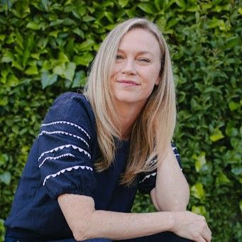 Blonde woman with straight hair resting her head on her hand, smiling softly against a leafy green background.