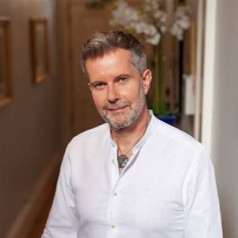 Middle-aged man with short gray hair and beard wearing a white shirt standing in a warmly lit hallway with blurred decor in the background.
