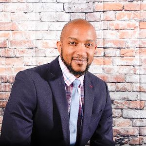 Man with a beard wearing a navy suit, plaid shirt, and light blue tie sitting in front of a brick wall.