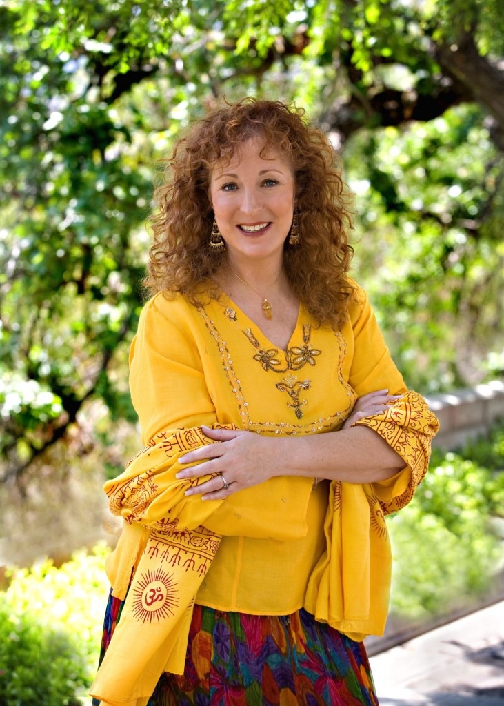 Smiling woman with curly red hair wearing a yellow embroidered top and colorful skirt standing outdoors with green foliage background.
