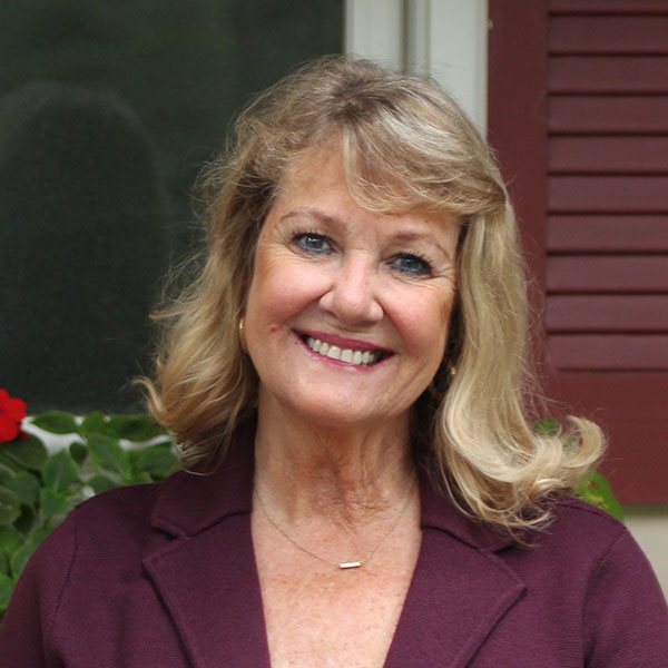 Smiling middle-aged woman with blonde hair wearing a maroon jacket and gold necklace, standing outdoors near plants and a window.