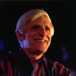 Older man with gray hair smiling, wearing a dark jacket against a dark background.