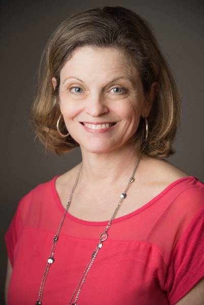 Smiling middle-aged woman with shoulder-length brown hair wearing a bright pink top, silver hoop earrings, and a long silver necklace against a plain dark background.