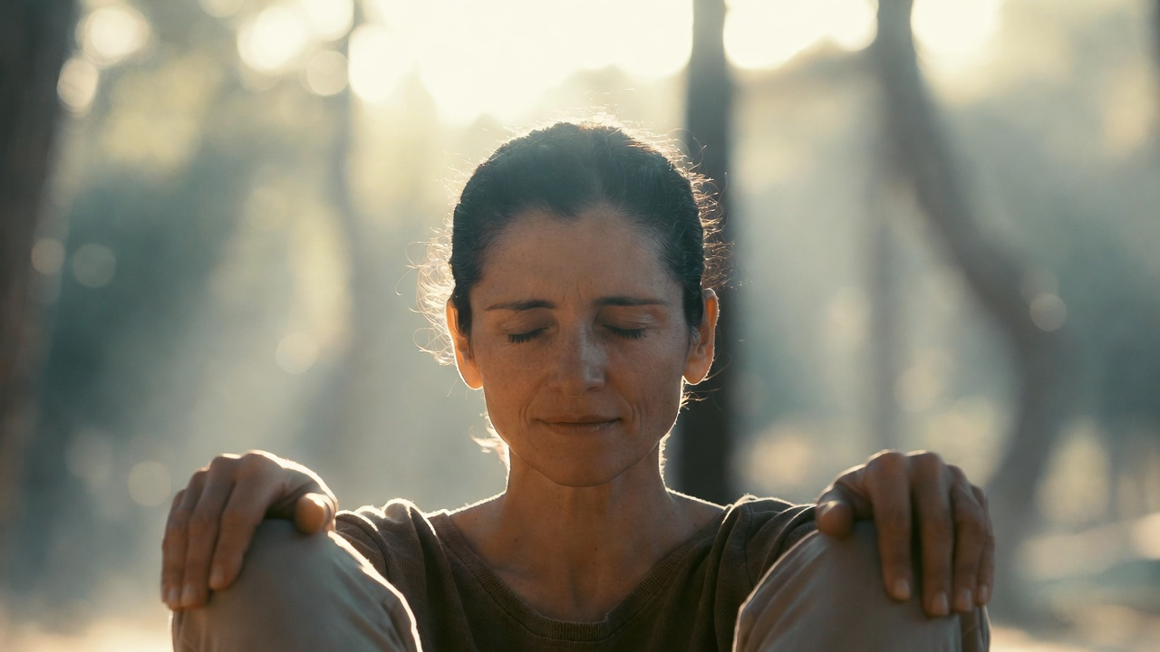 Woman with closed eyes sitting outdoors with knees drawn close, surrounded by soft sunlight and blurred trees.