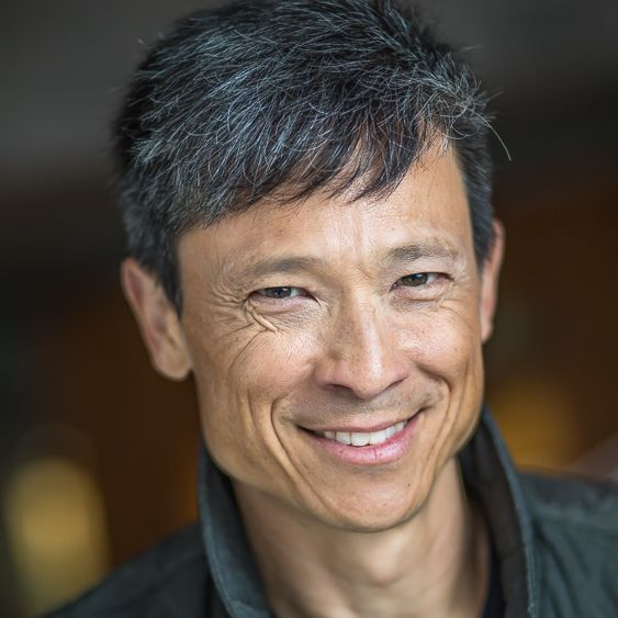 Smiling middle-aged man with short salt-and-pepper hair wearing a dark jacket against a blurred background.