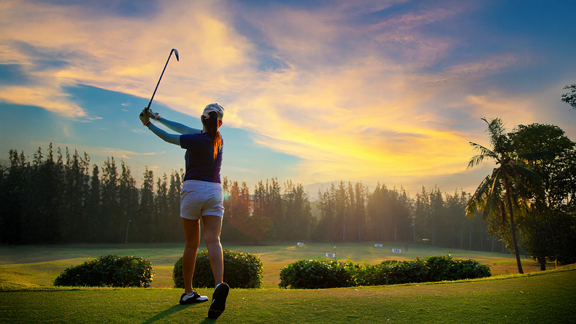 Golfer in shorts and cap swinging a golf club on a green field at sunset with trees in the background.