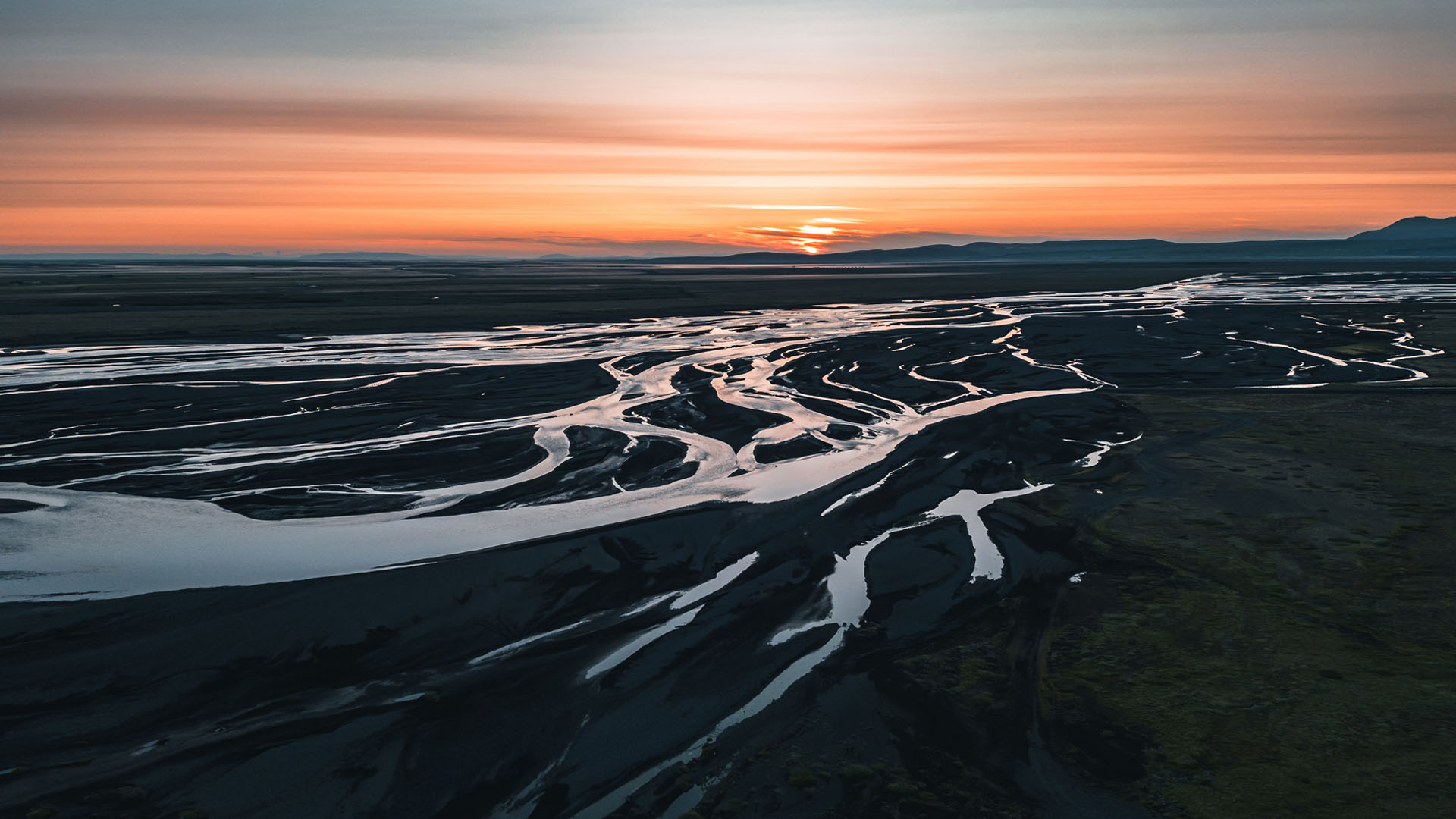 A braided river flowing through dark sandbeds at sunset with an orange and purple sky above distant mountains.