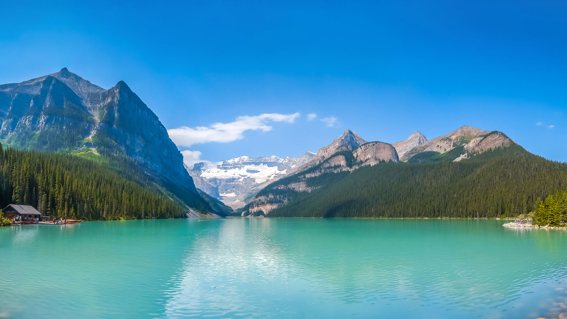 Turquoise lake surrounded by pine forests and towering mountains with snow-capped peaks under a clear blue sky.