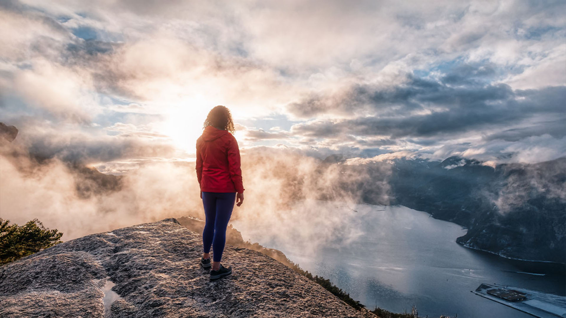 Person in red jacket and blue leggings standing on rocky cliff overlooking a foggy valley and body of water under a cloudy sky at sunrise.