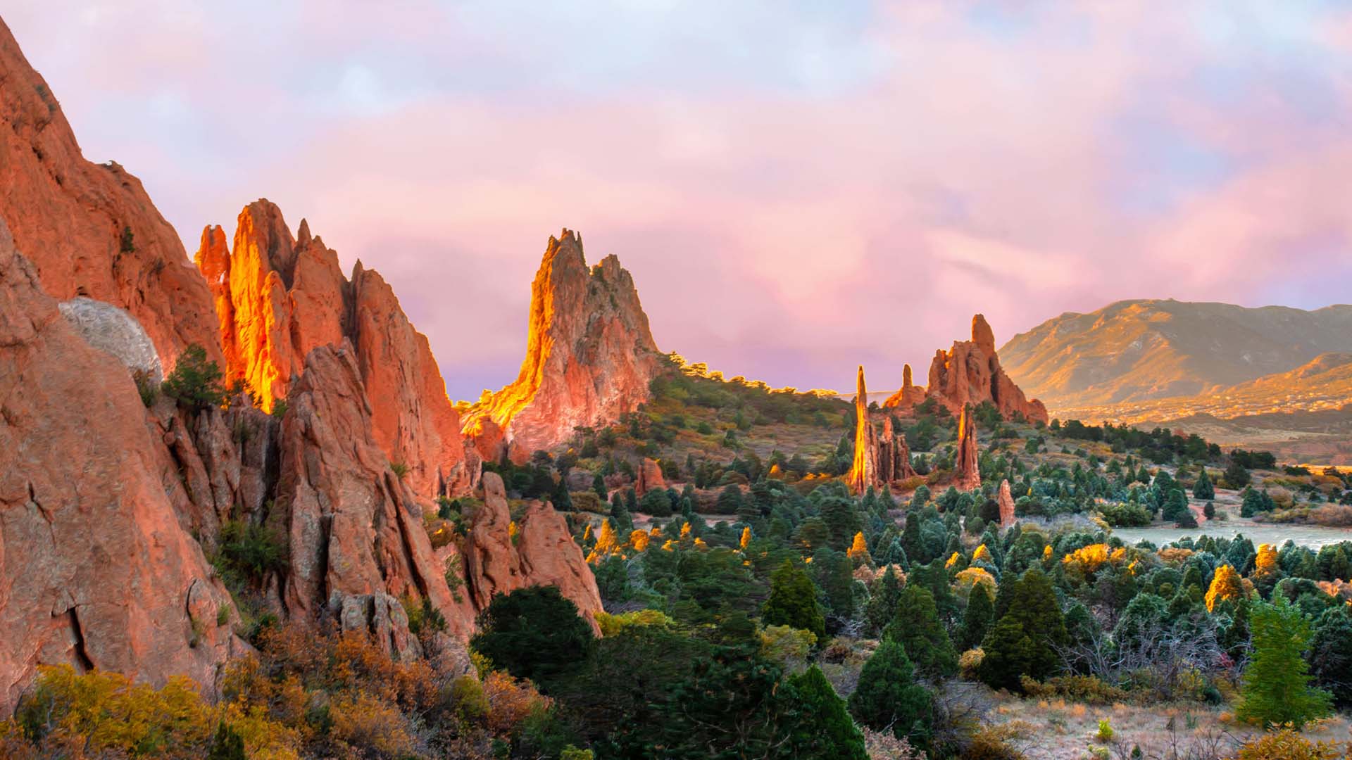 Sunlit red rock formations rising above a green forest under a pink and blue sky.